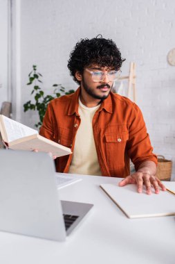 Indian copywriter in eyeglasses holding book near notebook and laptop at home 