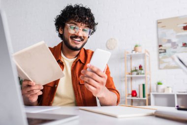 Smiling indian copywriter using smartphone and holding book near blurred laptop at home 