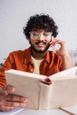 Indian copywriter talking on smartphone and holding blurred book at home 