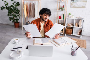Indian copywriter holding papers near devices and notebooks on table at home 