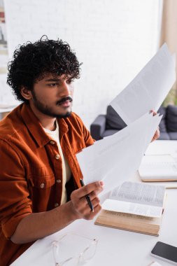 Curly indian copywriter working with papers near book and cellphone at home 