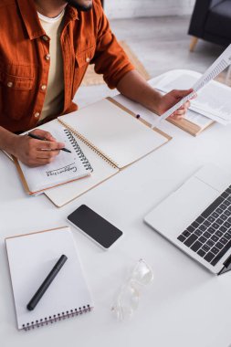 Cropped view of copywriter holding paper and writing on notebook near devices and eyeglasses at home 