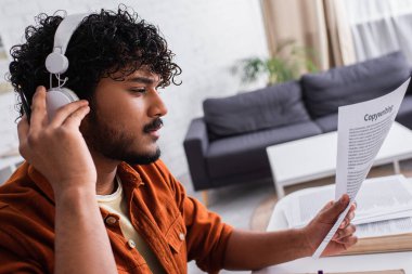 Indian copywriter in headphones holding paper at home 