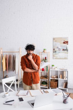 Indian copywriter in headphones standing near papers and gadgets on table at home 