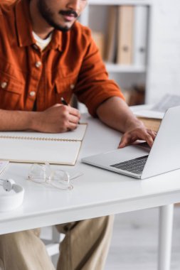 Cropped view of indian copywriter using laptop and writing on notebook near eyeglasses at home 