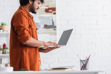 Side view of indian copywriter in eyeglasses holding laptop with blank screen at home 