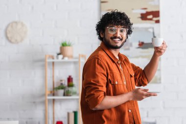 Smiling indian man in eyeglasses holding cup of coffee at home 