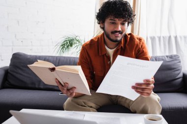 Smiling indian copywriter working with book and paper on couch at home 