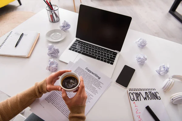 stock image Top view of freelancer holding cup of coffee near devices and papers with copywriting lettering on table 