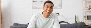 happy african american man looking at camera in living room, banner 