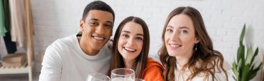cheerful women and african american man looking at camera in living room, banner 