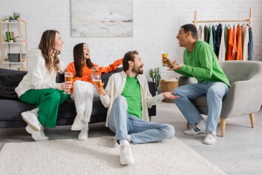 cheerful multicultural men and women holding glasses of beer while chatting in living room 