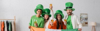 happy and multicultural friends in green hats holding glasses of beer and Irish flag while celebrating Saint Patrick Day, banner  clipart