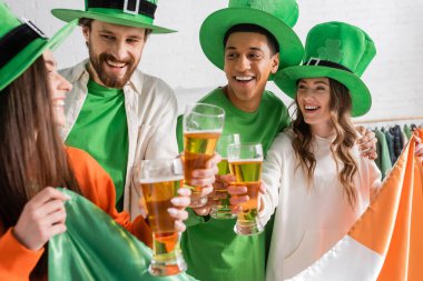 happy and interracial friends in green hats holding glasses of beer and Irish flag while celebrating Saint Patrick Day 