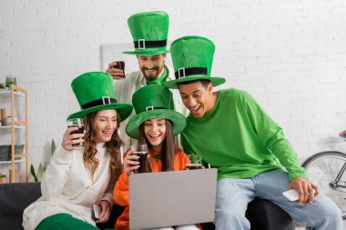 cheerful and interracial friends in green hats holding glasses of dark beer while looking at laptop 