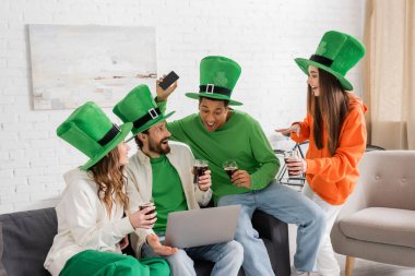 joyful and multiethnic friends in green hats holding glasses of dark beer during Saint Patrick Day celebration at home clipart