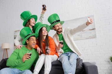 cheerful bearded man taking selfie with interracial friends holding glasses of dark beer on Saint Patrick Day clipart