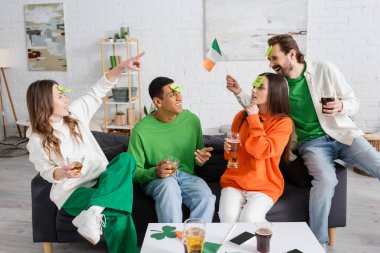 woman pointing at bearded man with sticky note on forehead holding Irish flag while playing guess who game with interracial friends  clipart
