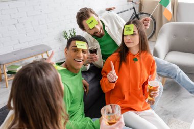 happy interracial friends with sticky notes on foreheads looking at woman while playing guess who game on Saint Patrick Day clipart