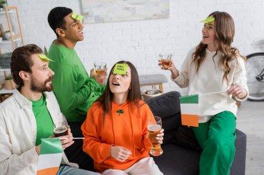 multiethnic friends with sticky notes on foreheads holding alcohol drinks and playing guess who game on Saint Patrick Day clipart
