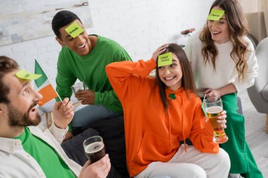 joyful multiethnic friends with sticky notes on foreheads holding alcohol drinks and playing guess who game on Saint Patrick Day clipart