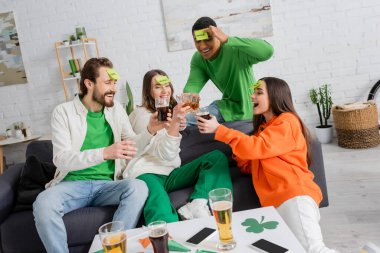 happy interracial friends with sticky notes clinking glasses of alcohol drinks and playing guess who game on Saint Patrick Day clipart