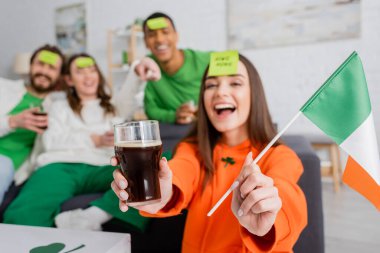 amazed woman with sticky note on forehead holding Irish flag and glass of beer near interracial friends on Saint Patrick Day