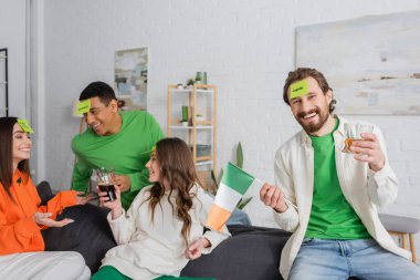 happy bearded man with apple word on sticky note holding Irish flag and drink near interracial friends on Saint Patrick Day
