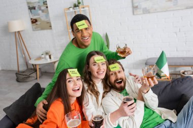 Cheerful interracial friends holding beer while playing who i am game at home 