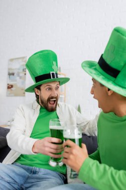Excited man in green hat holding beer near african american friend at home 
