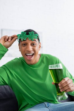 Excited african american man holding party sunglasses and holding beer during saint patrick day clipart