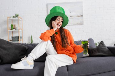 Positive woman in green hat holding glass of green beer on couch at home  clipart