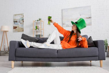 Side view of young woman in hat holding beer while celebrating saint patrick day clipart