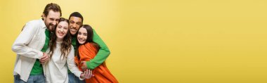Smiling interracial friends hugging and looking at camera isolated on yellow, banner 