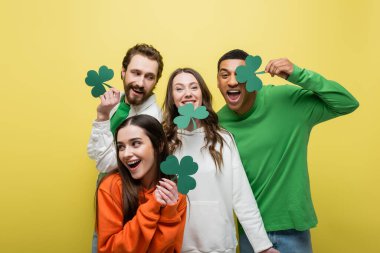 Cheerful interracial friends holding paper clover isolated on yellow 