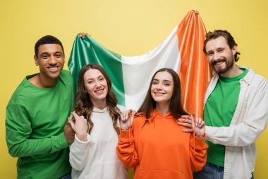 Smiling multiethnic men holding Irish flag near friends isolated on yellow  clipart