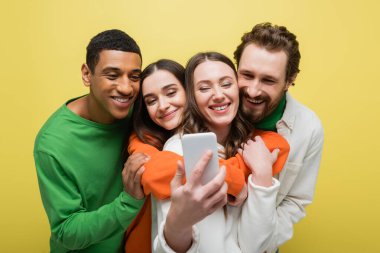 Positive interracial friends taking selfie on smartphone isolated on yellow 