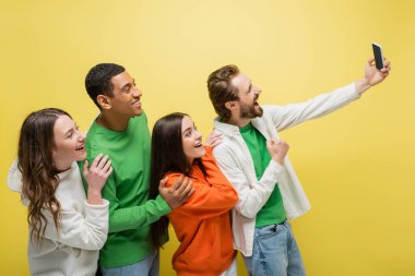 Positive man showing yes gesturing during video call near interracial friends isolated on yellow 