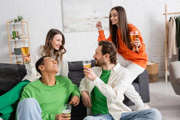 happy interracial group of friends holding glasses of beer while looking at each other in living room