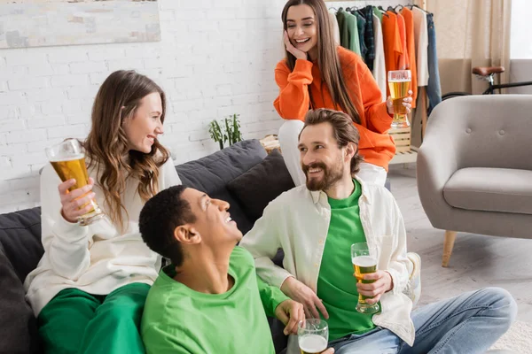 cheerful interracial group of friends holding glasses of beer while looking at each other in living room