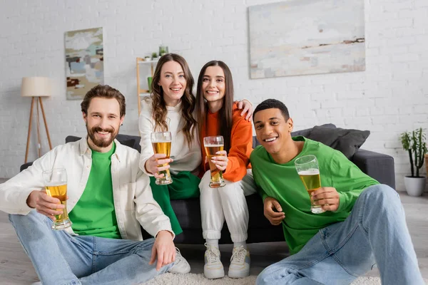 positive and interracial group of friends holding glasses of beer in living room