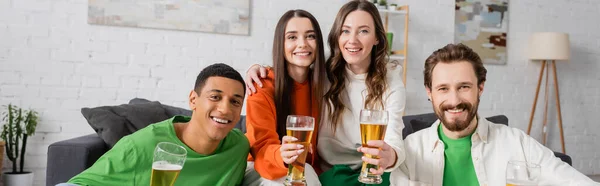 positive and interracial group of friends holding glasses of beer in living room, banner