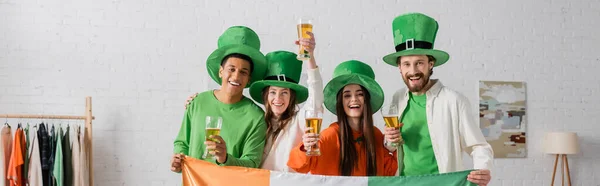 stock image happy and multicultural friends in green hats holding glasses of beer and Irish flag while celebrating Saint Patrick Day, banner 