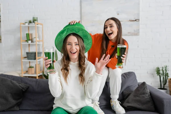Smiling woman in green hat looking at camera near friend with beer at home 