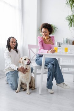 Smiling african american woman holding croissant near boyfriend and labrador in kitchen 