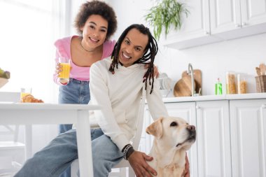 Positive african american couple looking at camera near labrador during breakfast in kitchen 