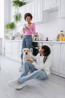 African american holding coffee near boyfriend petting labrador in kitchen 