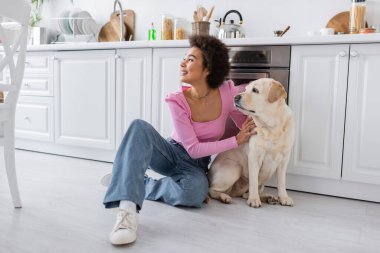 Cheerful african american woman petting labrador on floor at home 