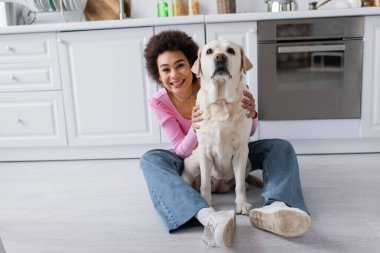 Young african american woman petting labrador on floor in kitchen 