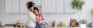 Smiling african american couple with orange juice hugging in kitchen, banner 